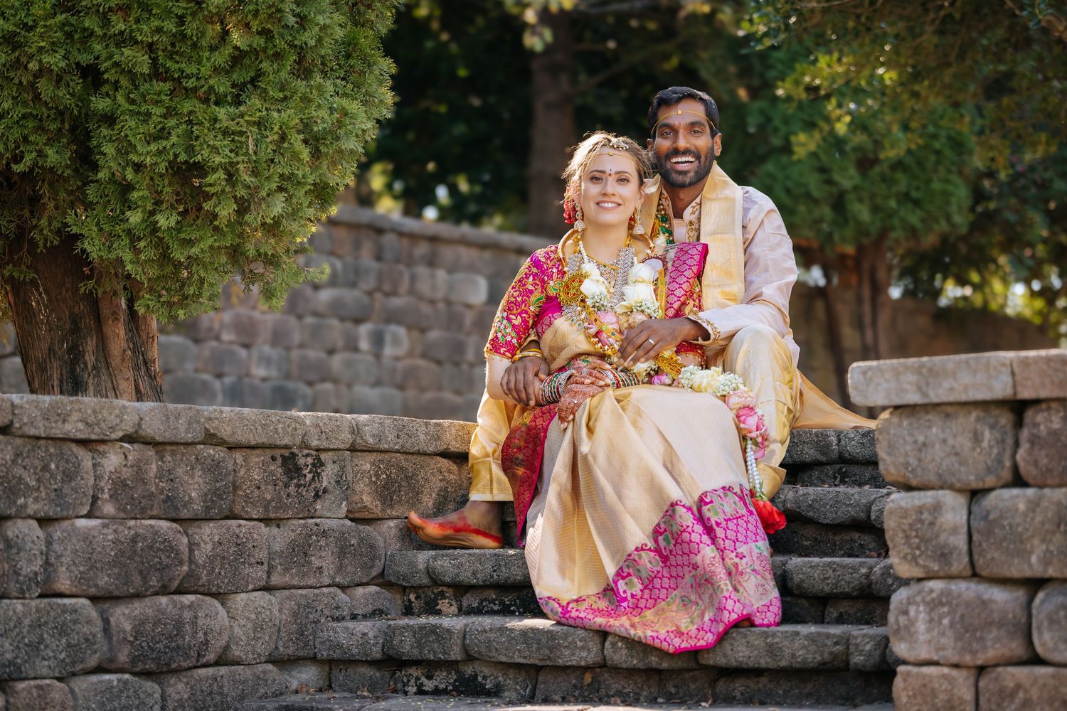 Smiling couple in traditional colorful Indian wedding attire with floral garlands sitting together on stone steps outdoors.