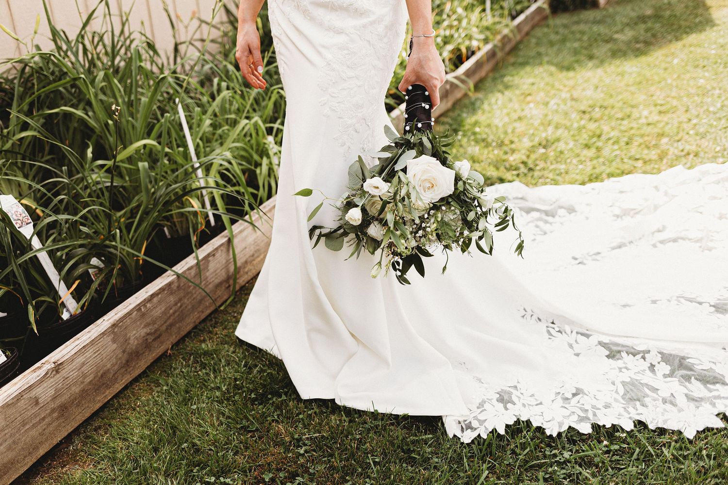 Detail of a bride’s lace wedding dress train spread across the grass as she holds her bouquet.