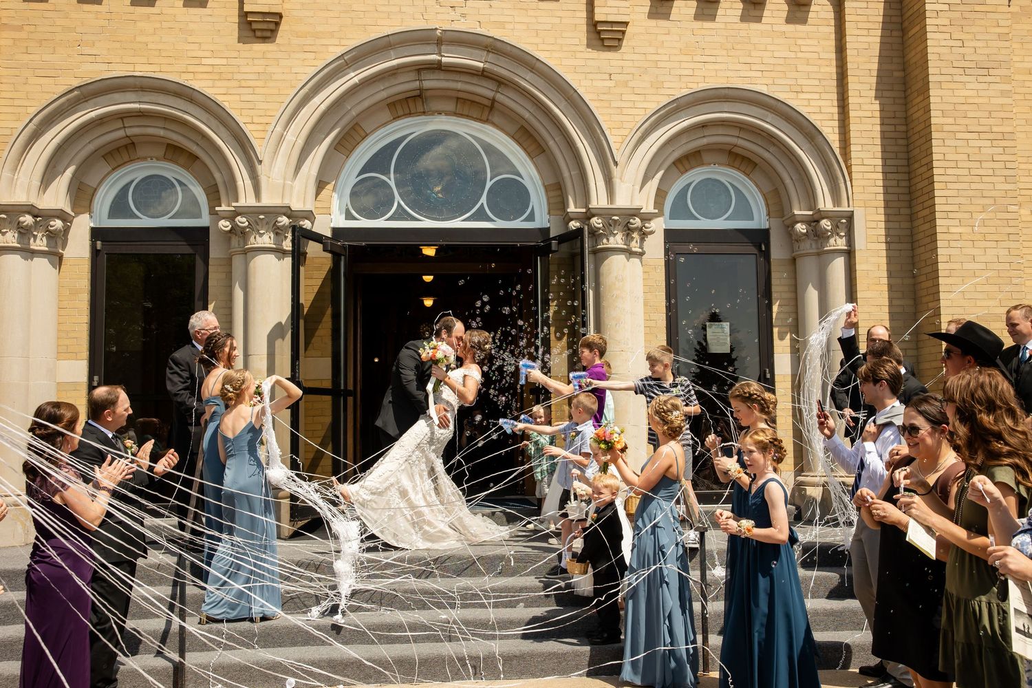 Bride and groom kiss on church steps as wedding guests in colorful dresses toss rice and cheer outside a historic brick building.
