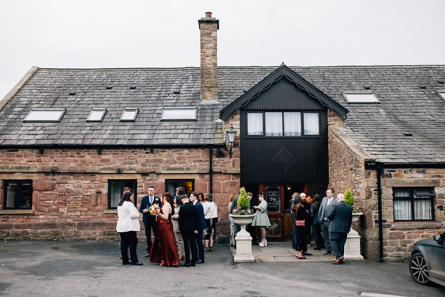 Stone countryside wedding venue with large glass-fronted barn, neatly mowed lawn, and stone steps framed by blurred yellow-green foliage.