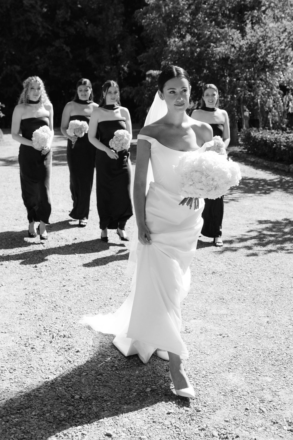 Bride in white gown leads bridesmaids in black dresses along a garden path, all holding bouquets, in black and white photo.