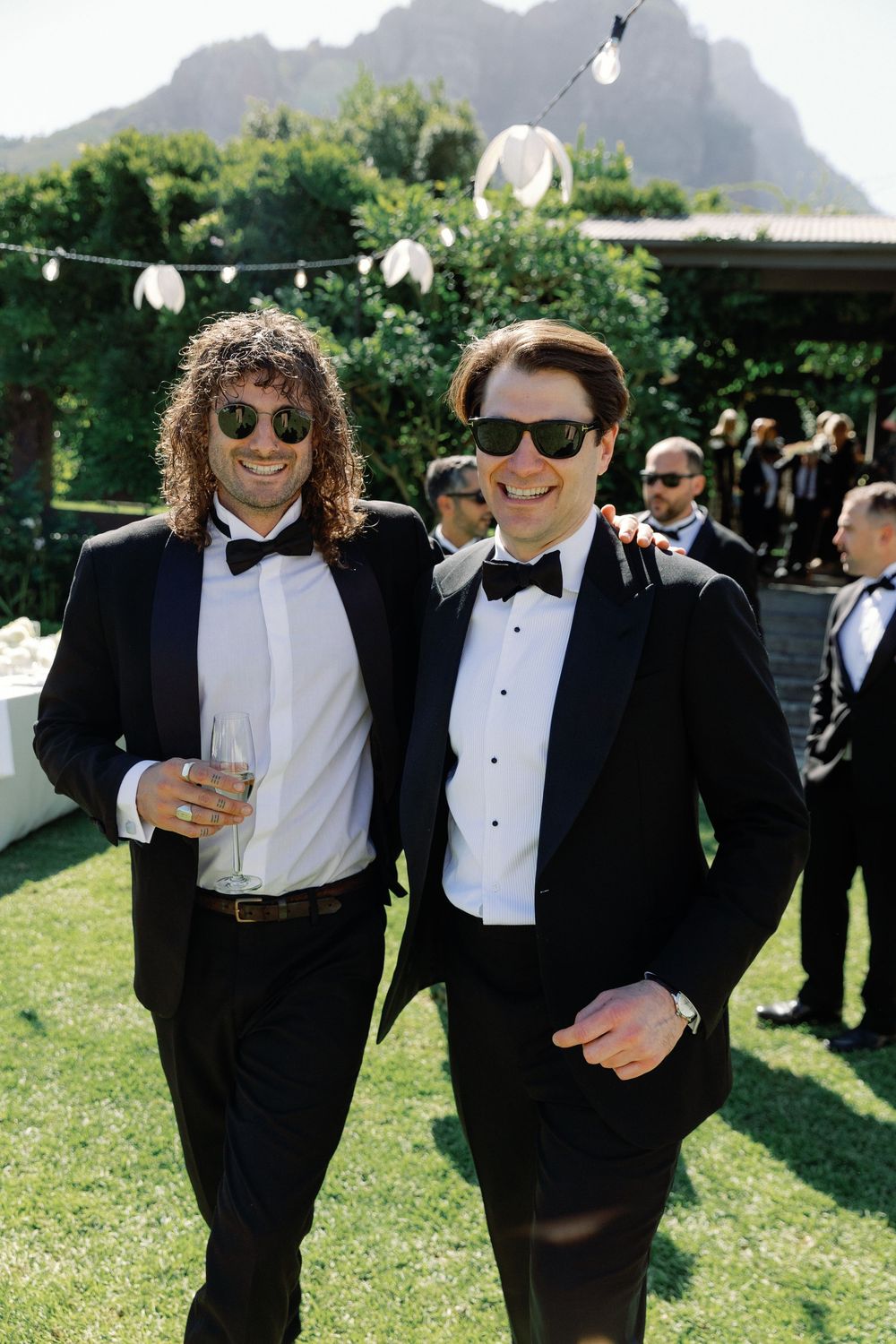 Two men in black tuxedos and sunglasses smile together at an outdoor wedding reception with mountain views.