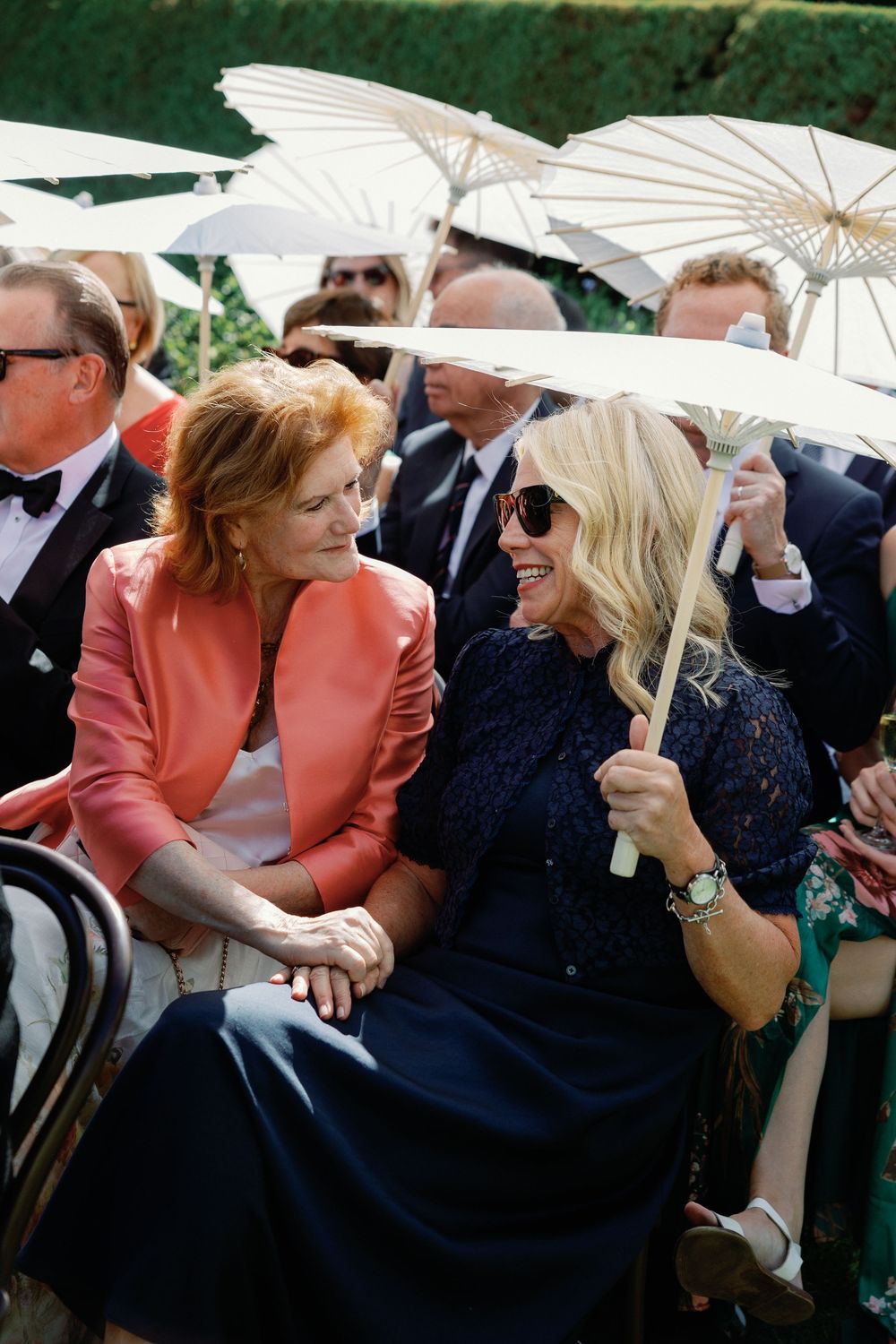 Two women laugh together at an outdoor wedding, one in coral blazer, other in navy lace dress holding white parasol.