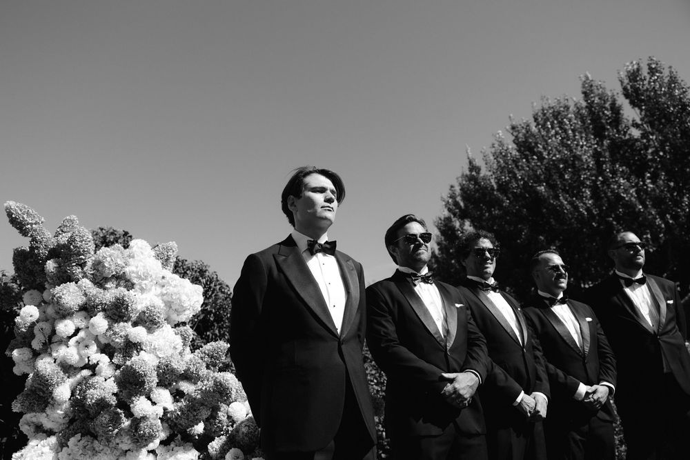 Black and white photo of groomsmen in tuxedos standing outdoors near floral arrangements at a wedding ceremony.