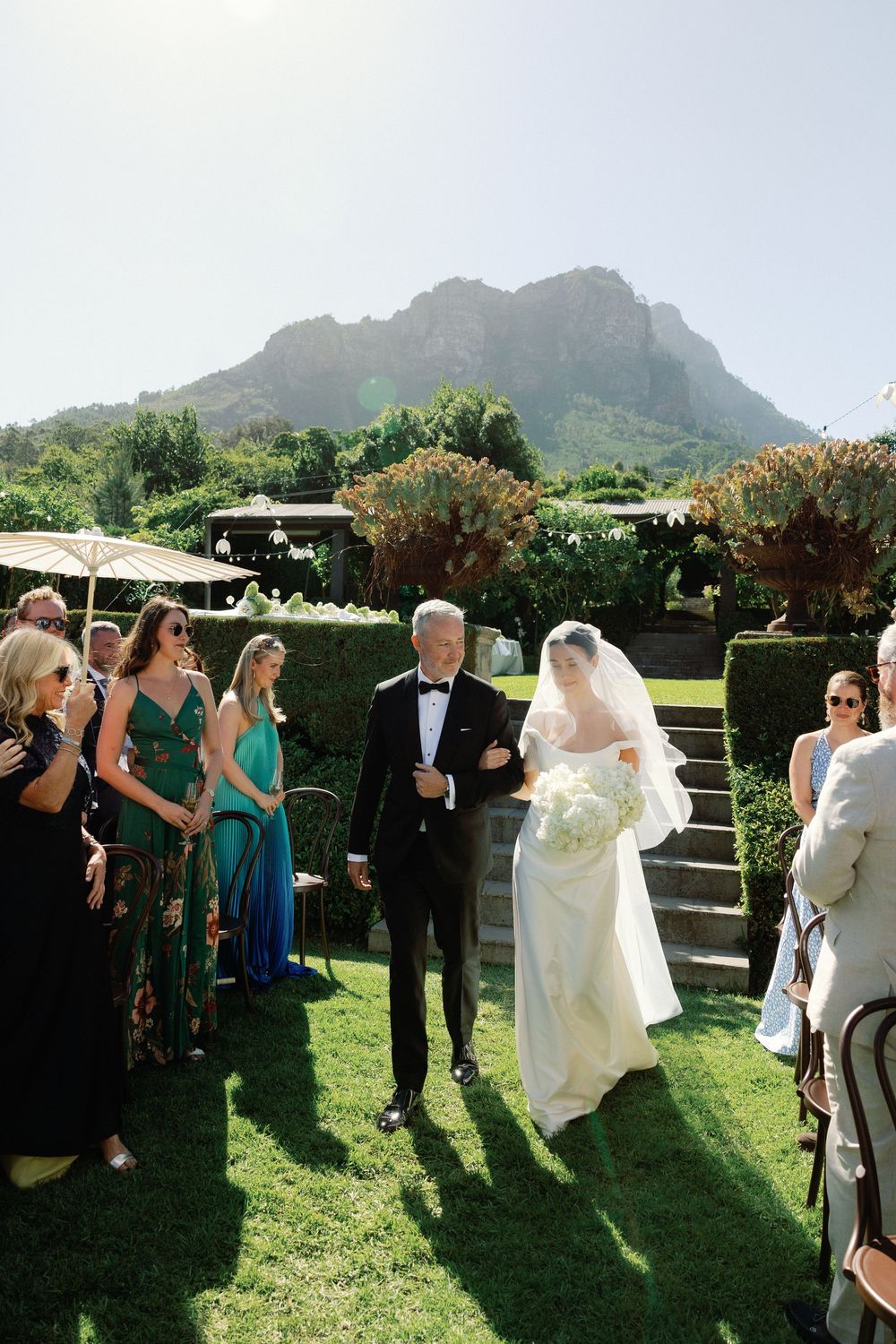 Bride walks down outdoor aisle with father in tuxedo, guests in green gowns, mountain backdrop at garden wedding.