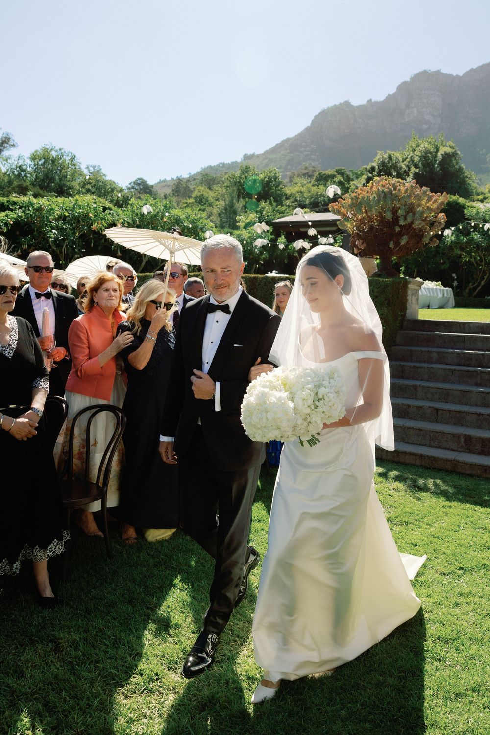 Bride in white gown and veil walks down outdoor aisle with escort, holding white bouquet, mountains in background.