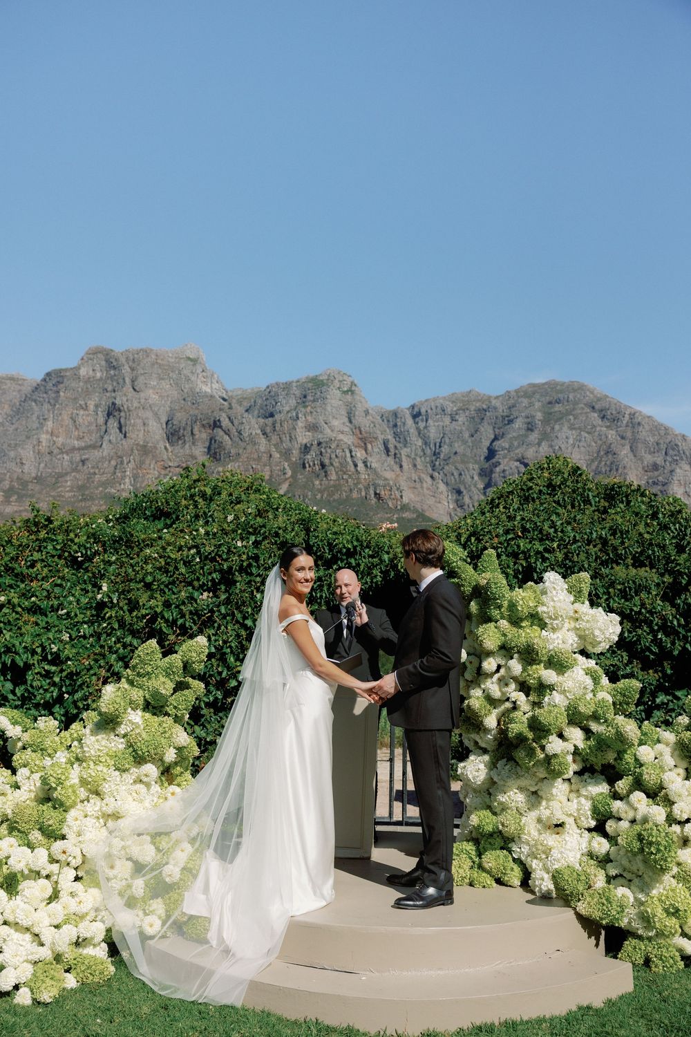 Bride and groom exchange vows at outdoor wedding ceremony with white hydrangeas and mountain backdrop under clear blue sky.