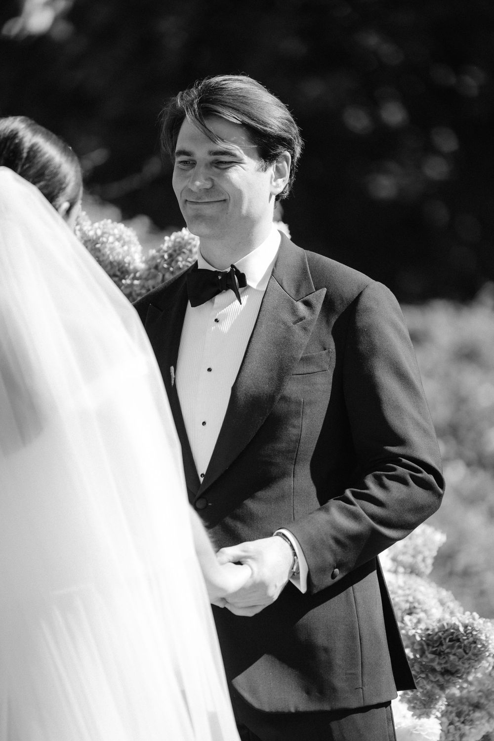 Groom in tuxedo smiling at bride during outdoor wedding ceremony, black and white photo.