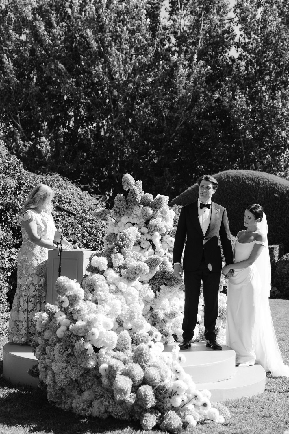 Black and white outdoor wedding ceremony with couple at floral altar and officiant at podium.