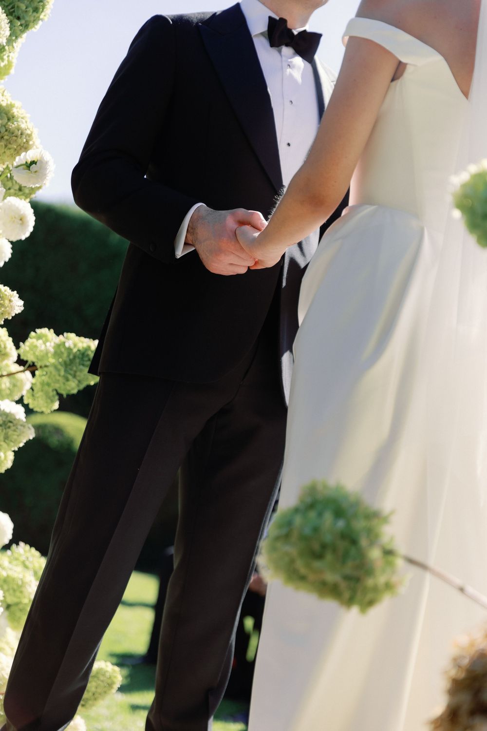 Bride and groom holding hands outdoors, groom in black tuxedo, bride in white off-shoulder gown, surrounded by greenery.