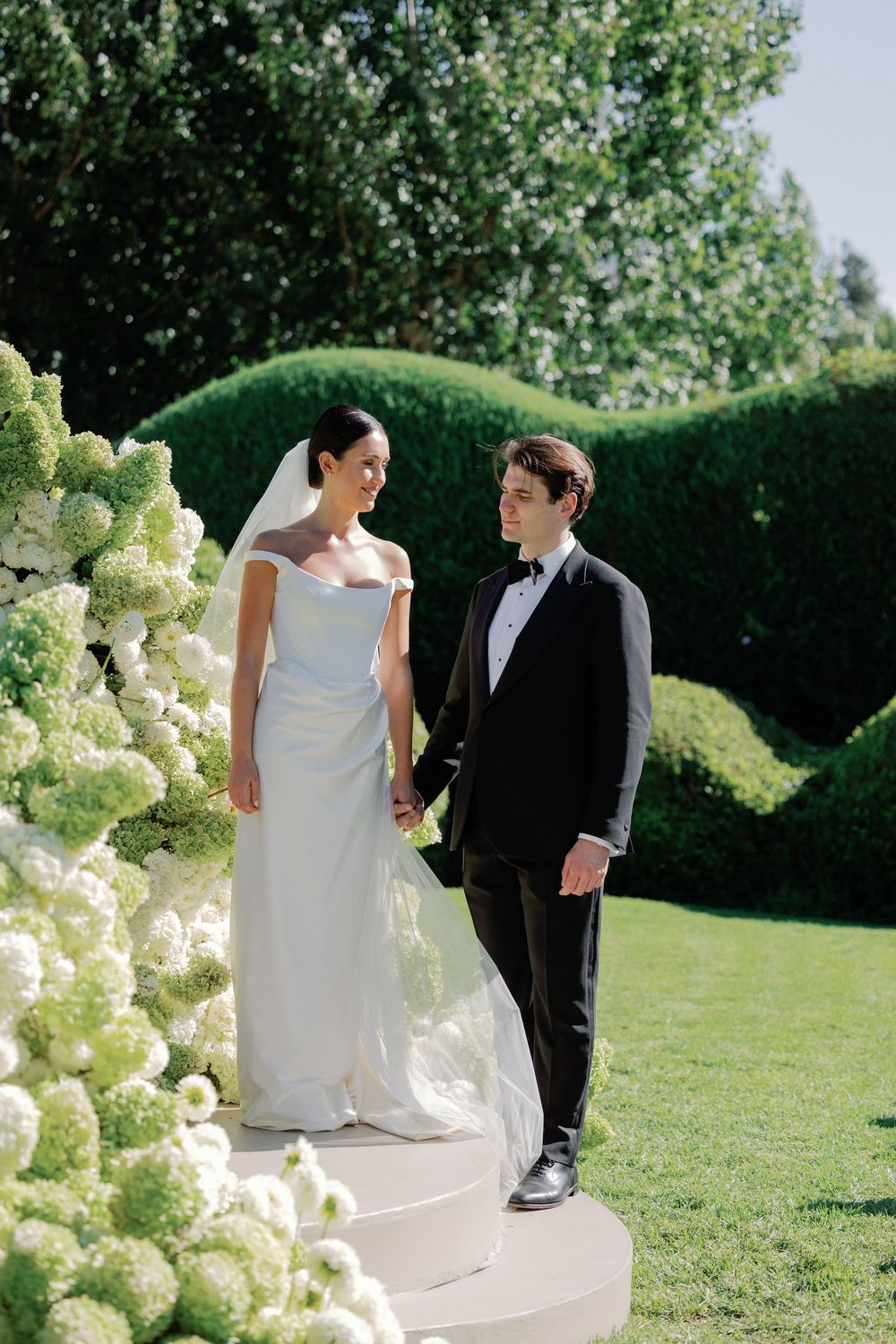 Bride in white off-shoulder gown and groom in black tuxedo hold hands in lush garden with hydrangeas.
