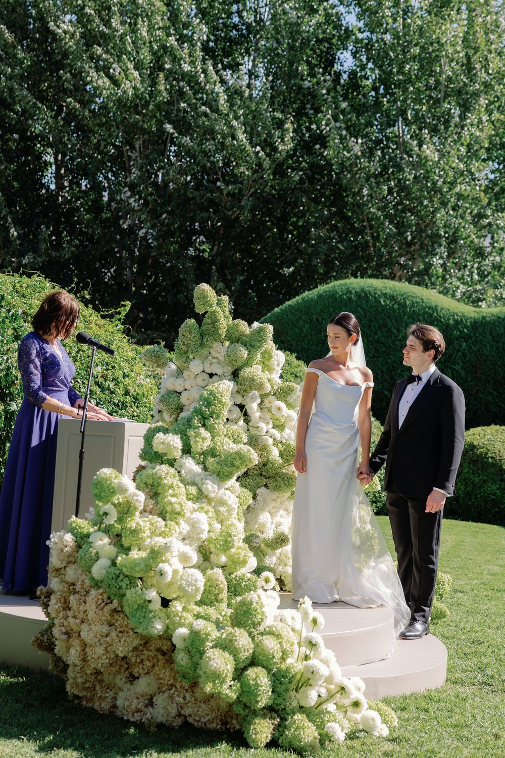 Bride and groom stand at outdoor wedding altar adorned with lush white hydrangea floral arrangements while officiant reads ceremony.