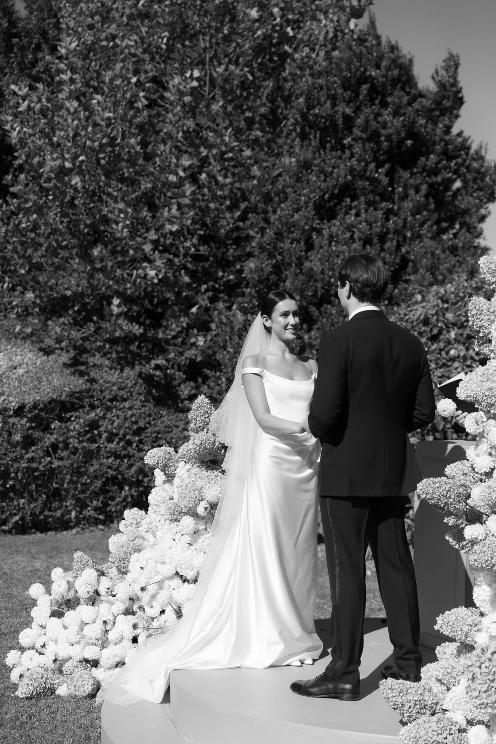 Black and white photo of bride and groom exchanging vows outdoors surrounded by hydrangea floral arrangements.