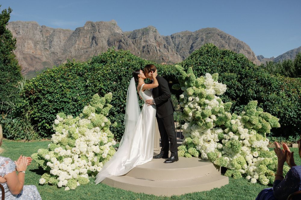 Couple shares first kiss at outdoor wedding ceremony with white hydrangea floral arch and mountain backdrop.