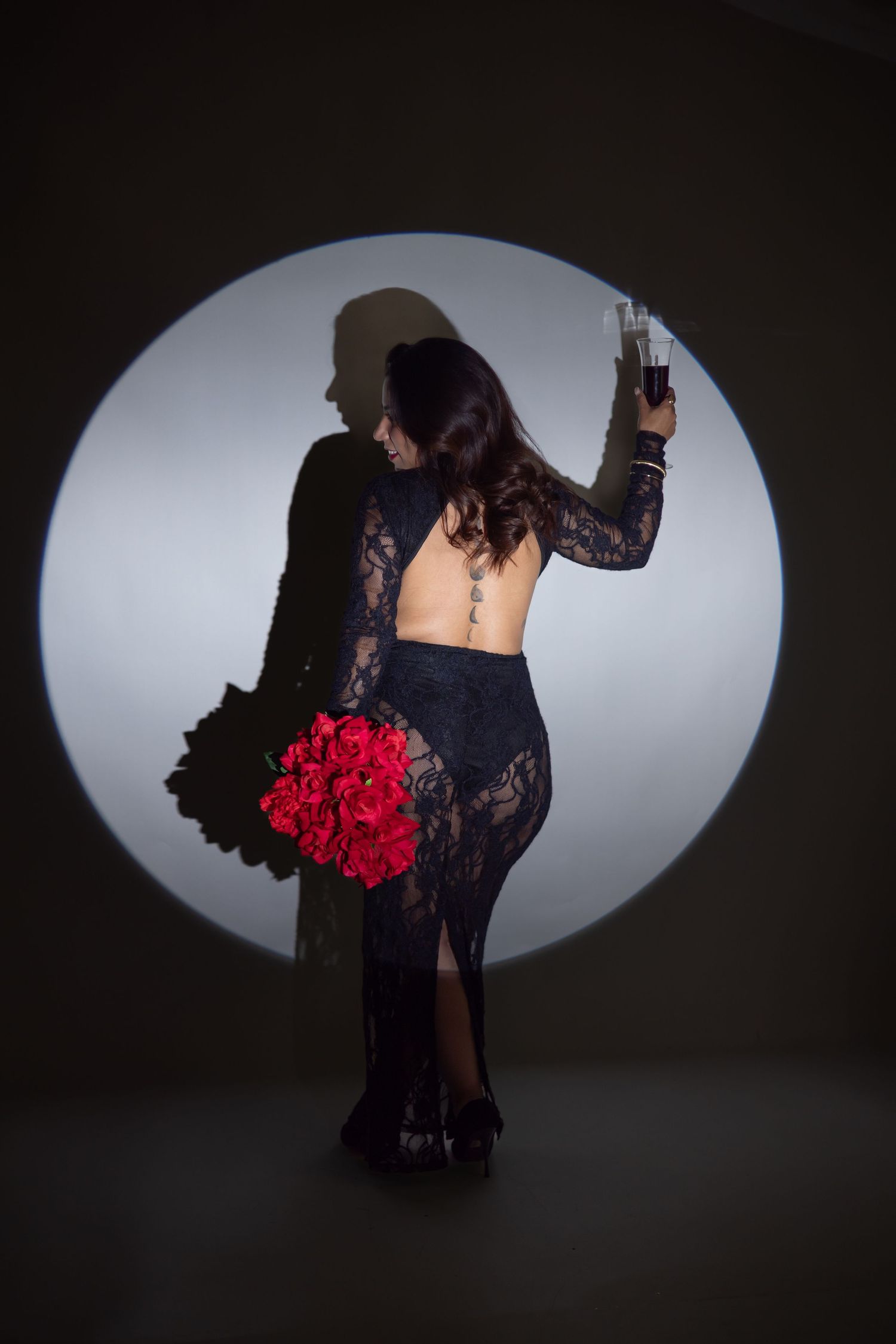 Woman in backless black lace gown holding red roses bouquet and champagne glass, posing before circular spotlight.