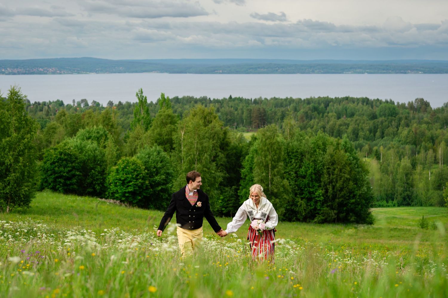 Couple in traditional attire holding hands walking through a lush green meadow with forest and lake in background.