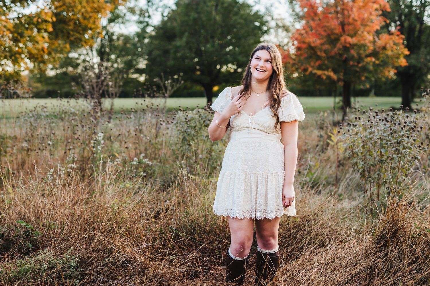 Young woman in white lace dress and brown boots standing in autumn field with colorful foliage background.