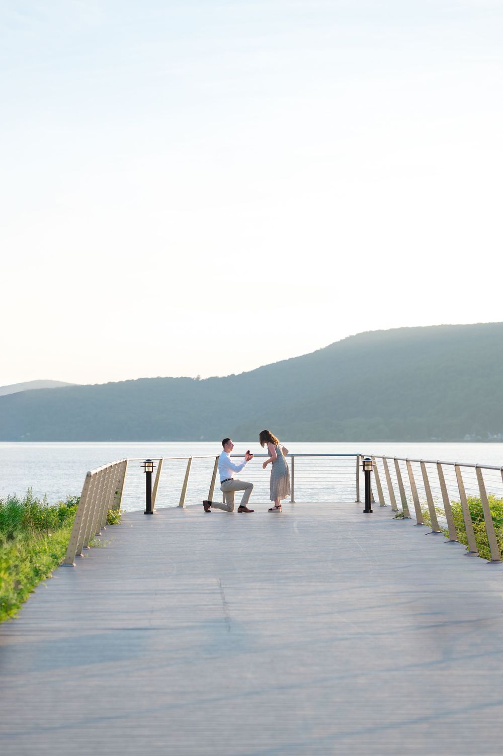 Man proposes on a scenic waterfront boardwalk with mountains and calm lake in the background at sunset.