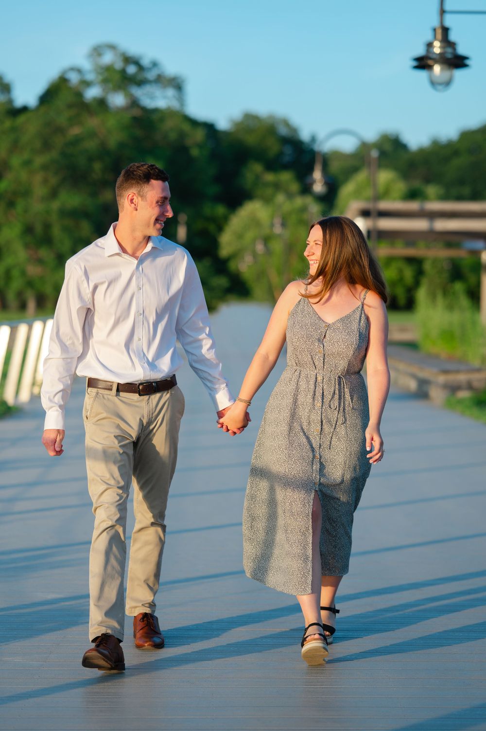 Couple holding hands and smiling while walking on a wooden boardwalk surrounded by lush greenery on a sunny day.