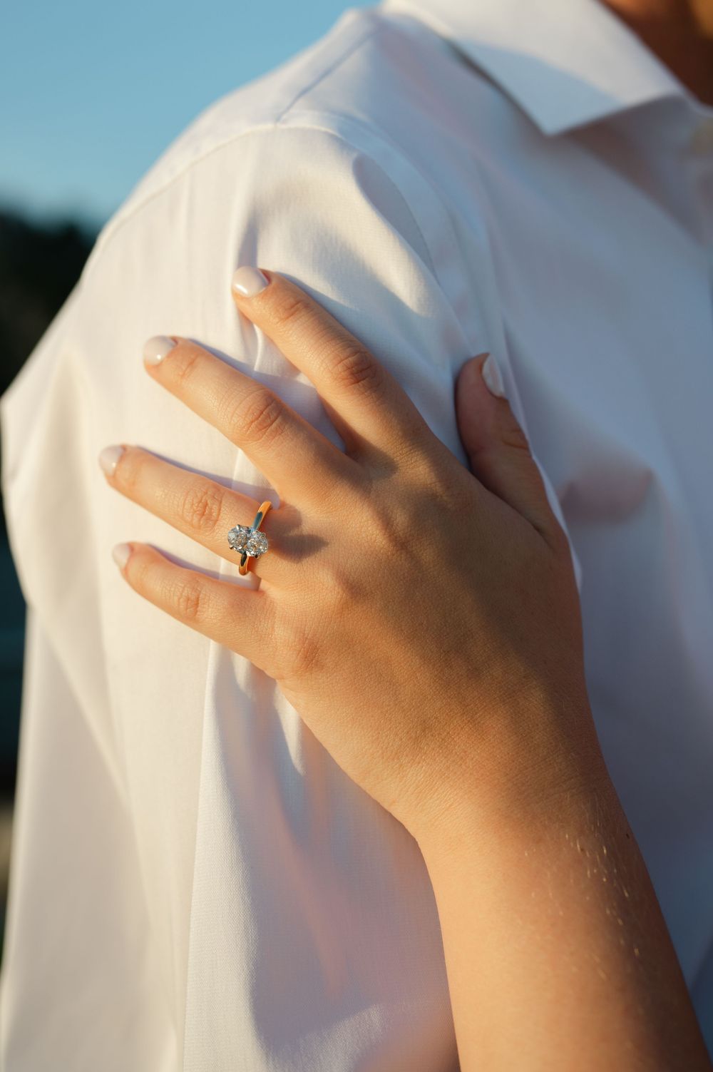 Close-up of a hand with an elegant diamond engagement ring resting on a white shirt shoulder in golden sunlight.
