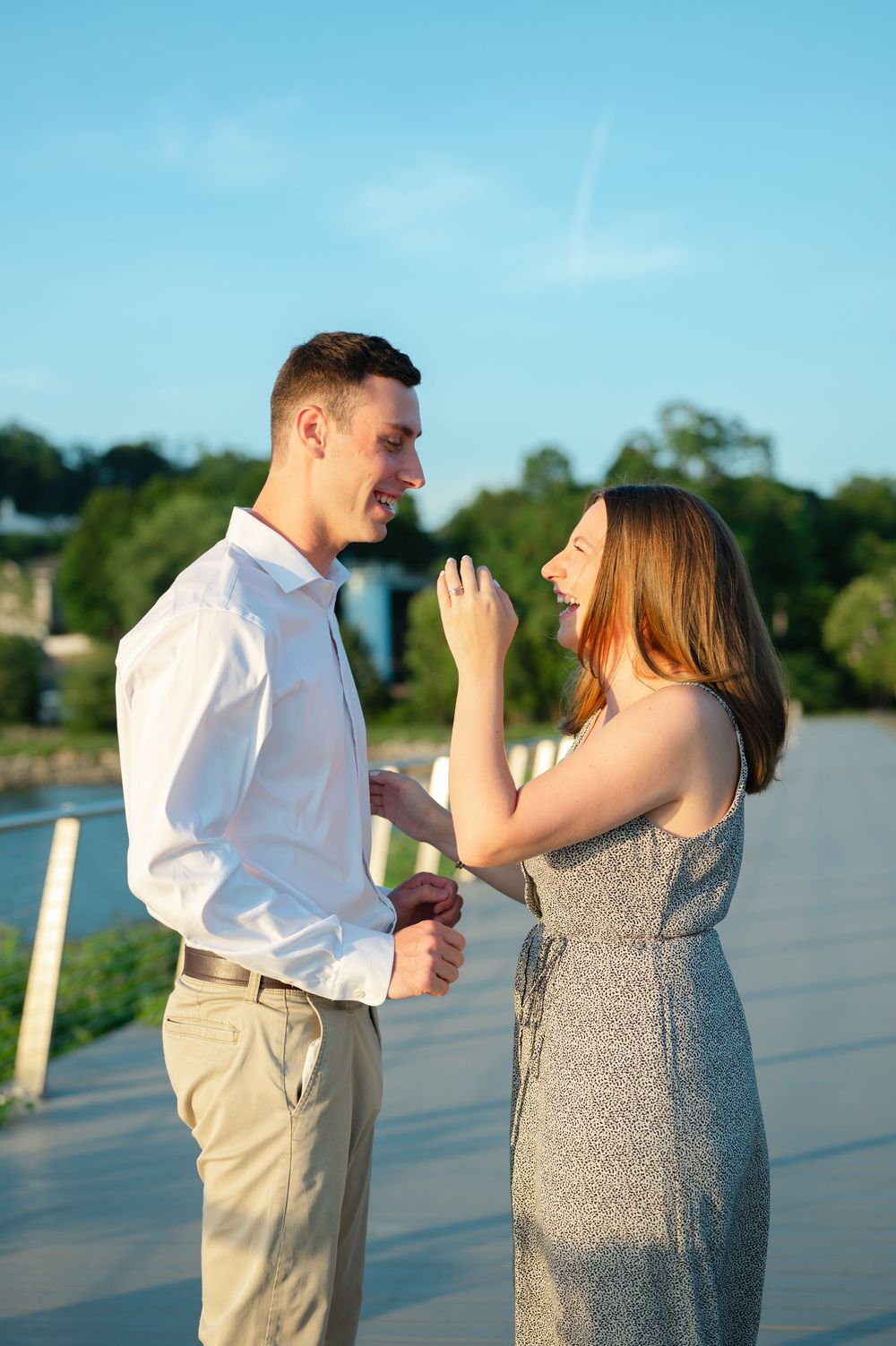 Happy couple sharing a romantic moment on a sunny dock near a river, woman showing her engagement ring.