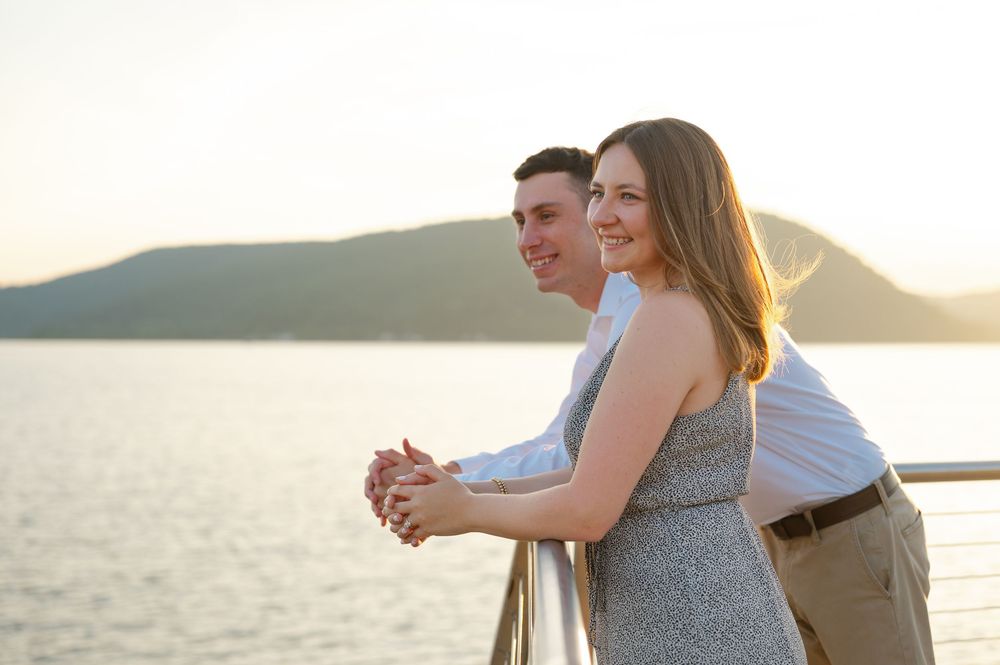 Couple smiling and holding hands on a waterfront railing at sunset with mountain views.