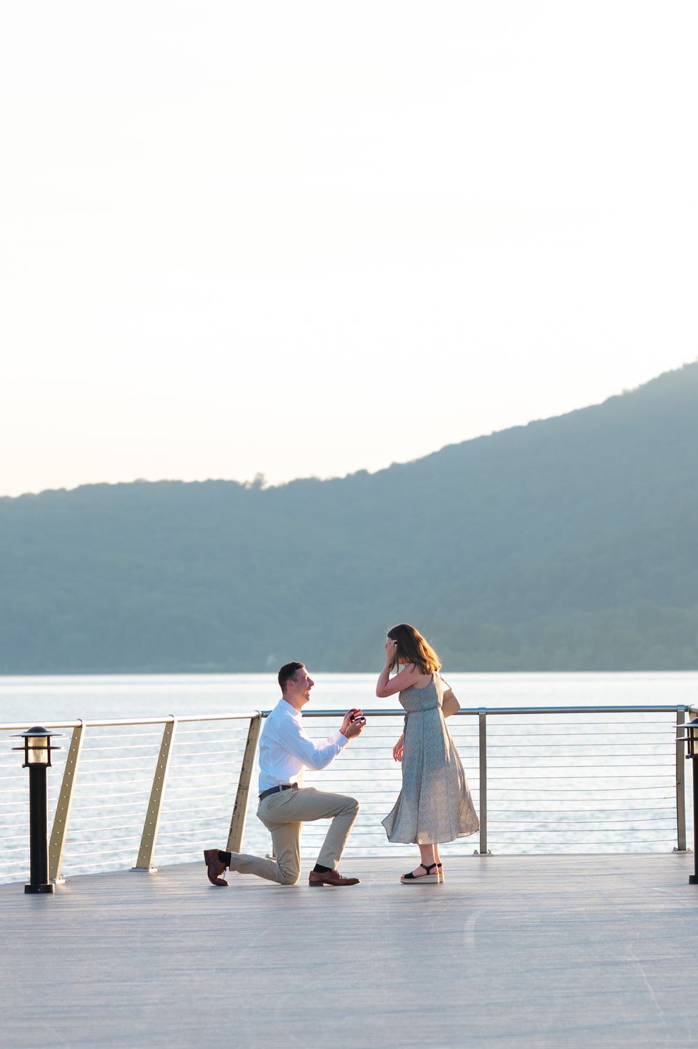 Man proposes to woman on a scenic waterfront deck with mountain and lake views at sunset.