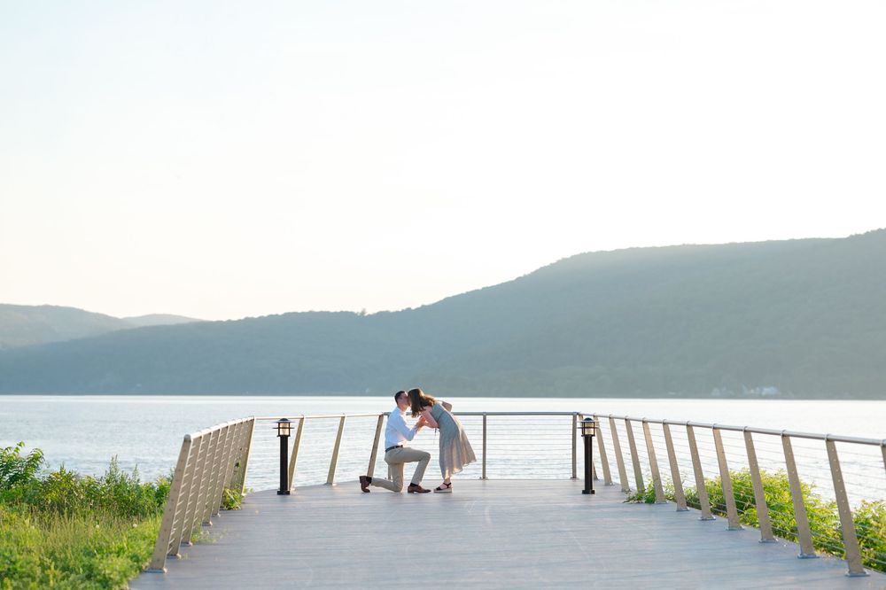 Couple sharing a romantic marriage proposal moment on a scenic wooden deck overlooking a calm lake with mountain views.