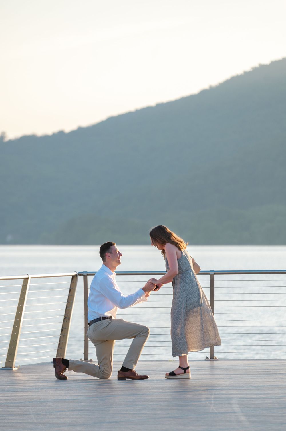 Man proposing on one knee to woman on waterfront deck with mountain lake backdrop at sunset.