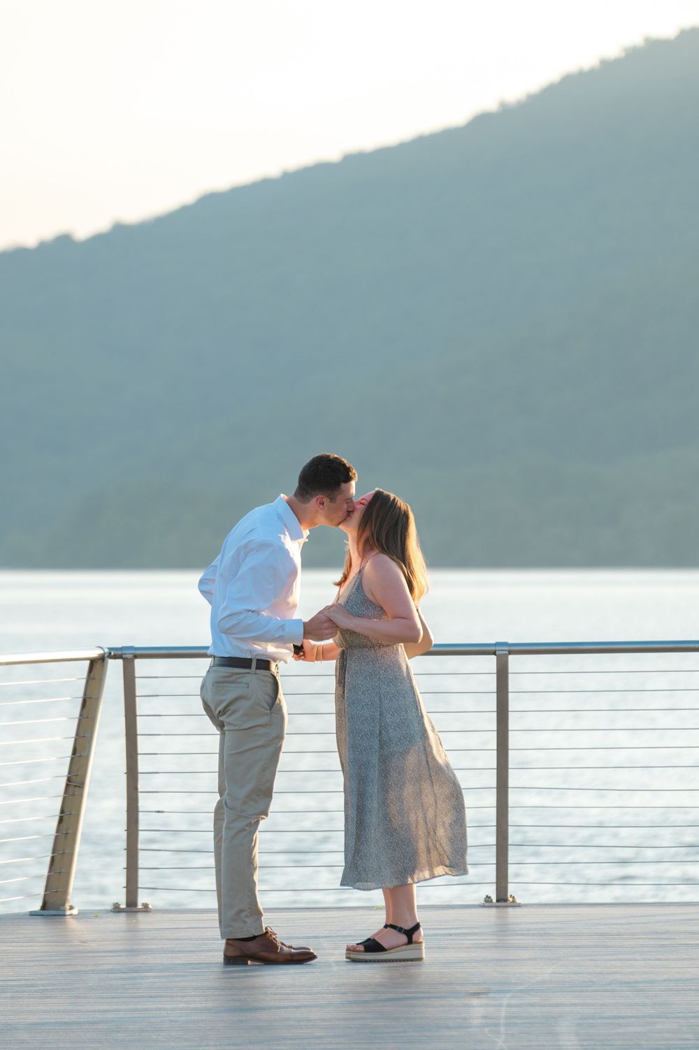 Couple kissing on a waterfront deck at sunset with mountain and lake views in the background.