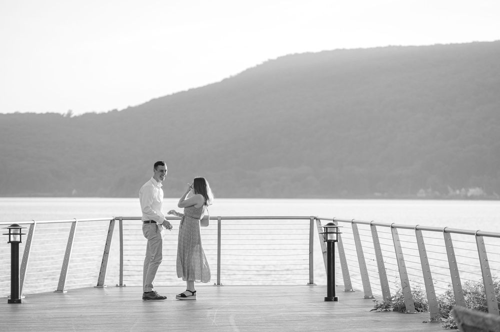 Couple standing on a waterfront deck with mountain views in a romantic black and white engagement photo.