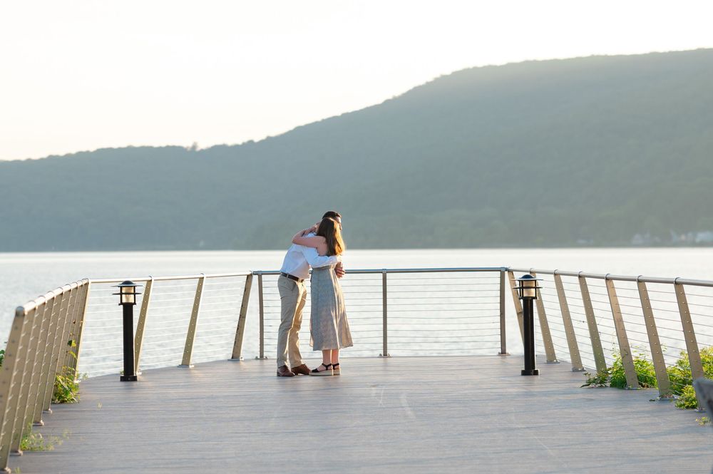 Couple embracing on a waterfront deck overlooking a serene lake with green hills at sunset.