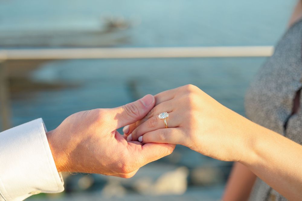 Person placing a diamond engagement ring on a partner's finger outdoors near water.