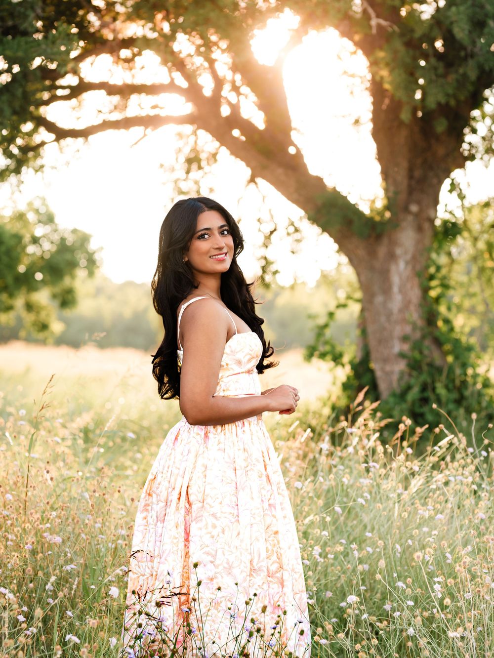Young woman in floral dress standing in sunlit meadow with tall grass and oak tree golden hour portrait.