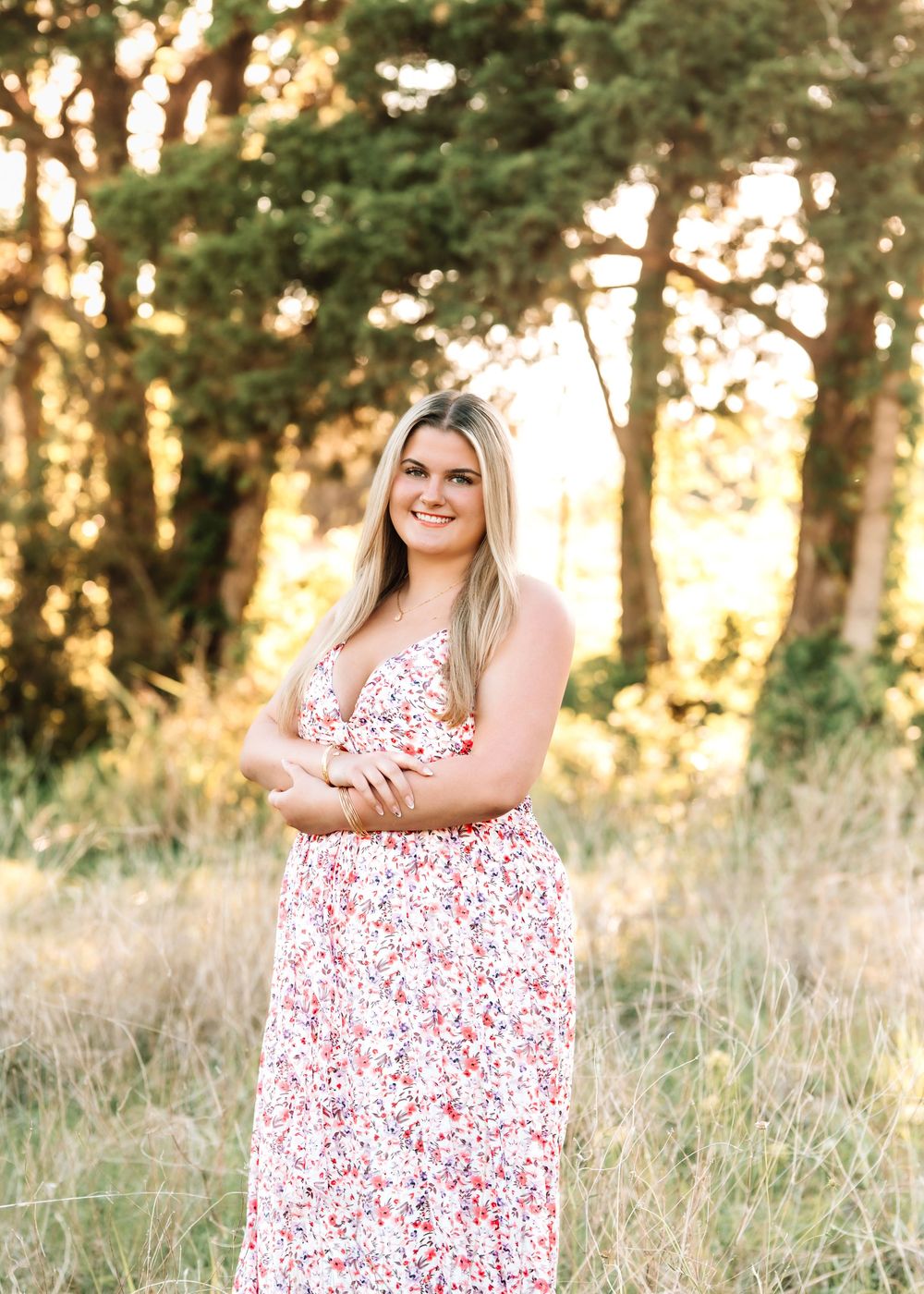 Young woman in floral maxi dress smiling outdoors among trees and golden grass in a nature setting.