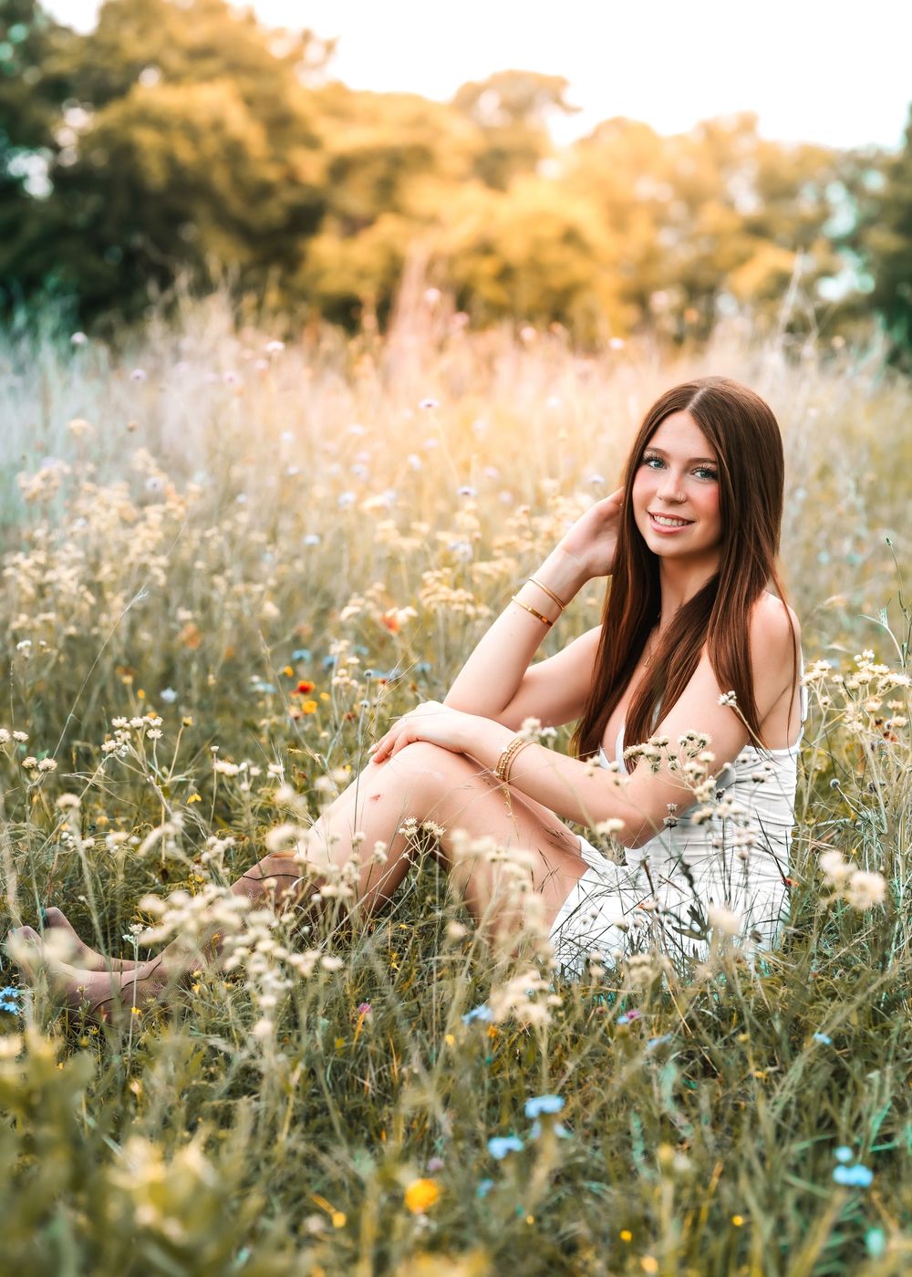 Young woman in floral dress and boots sitting in a wildflower meadow during golden hour sunset.