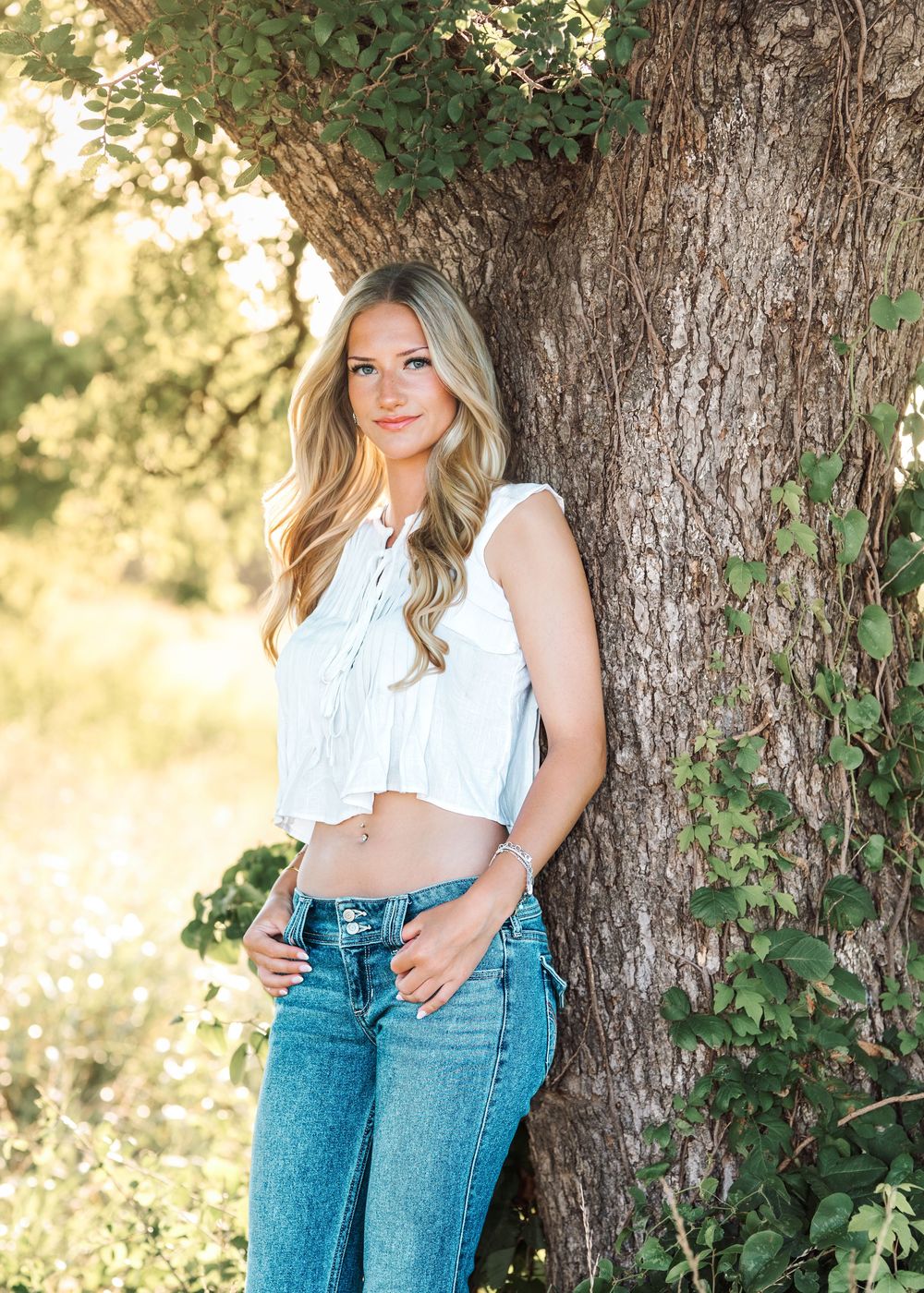 Young woman in white crop top and jeans leaning against a large tree outdoors in natural sunlight.