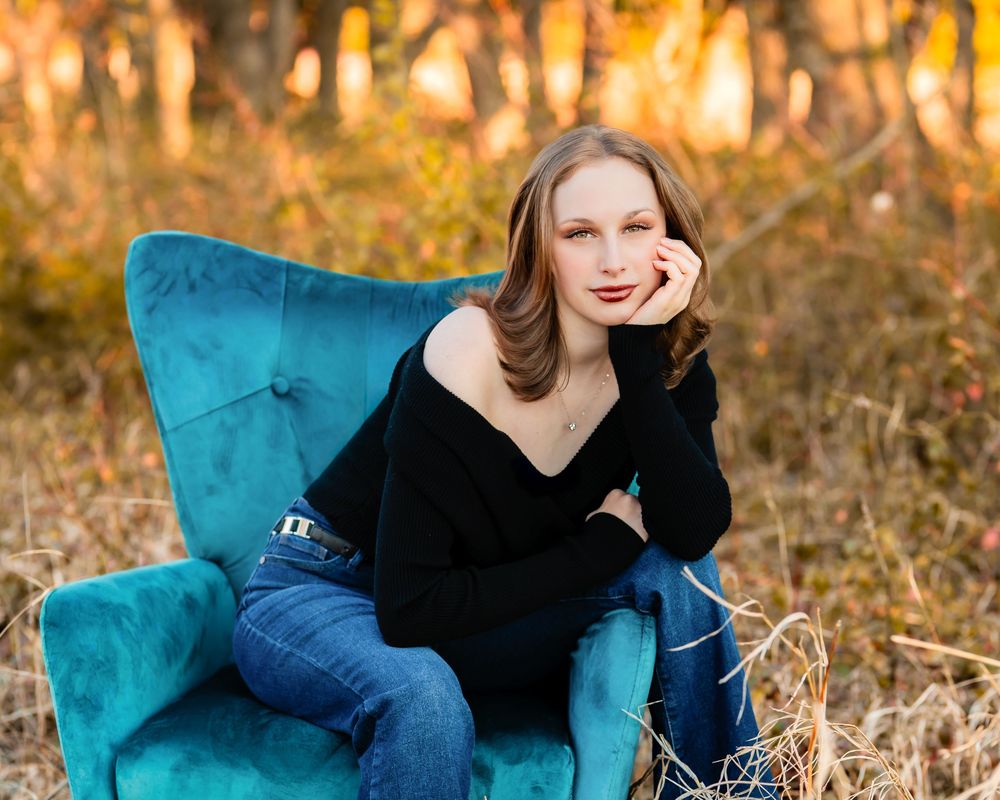 Young woman in black sweater and jeans poses gracefully on a teal velvet chair outdoors in golden autumn light.