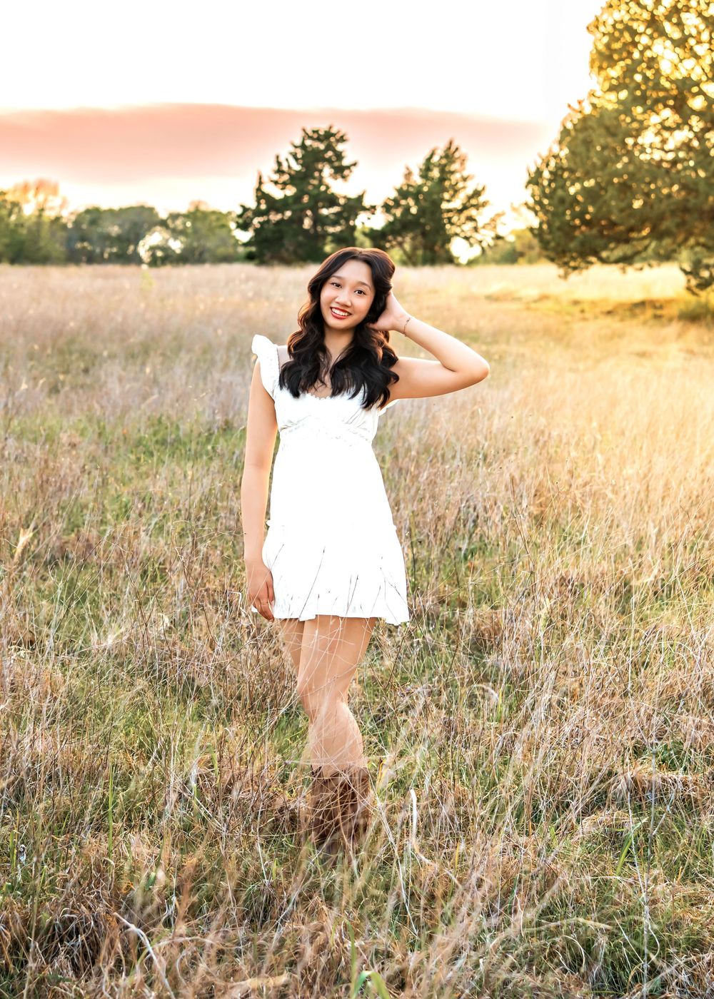 Young woman in white dress and brown boots smiling in golden field at sunset.