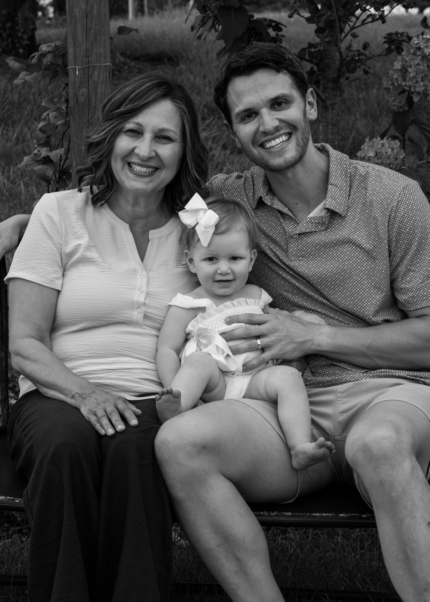 Black and white family portrait of a smiling grandmother, baby girl with bow, and young man seated outdoors.