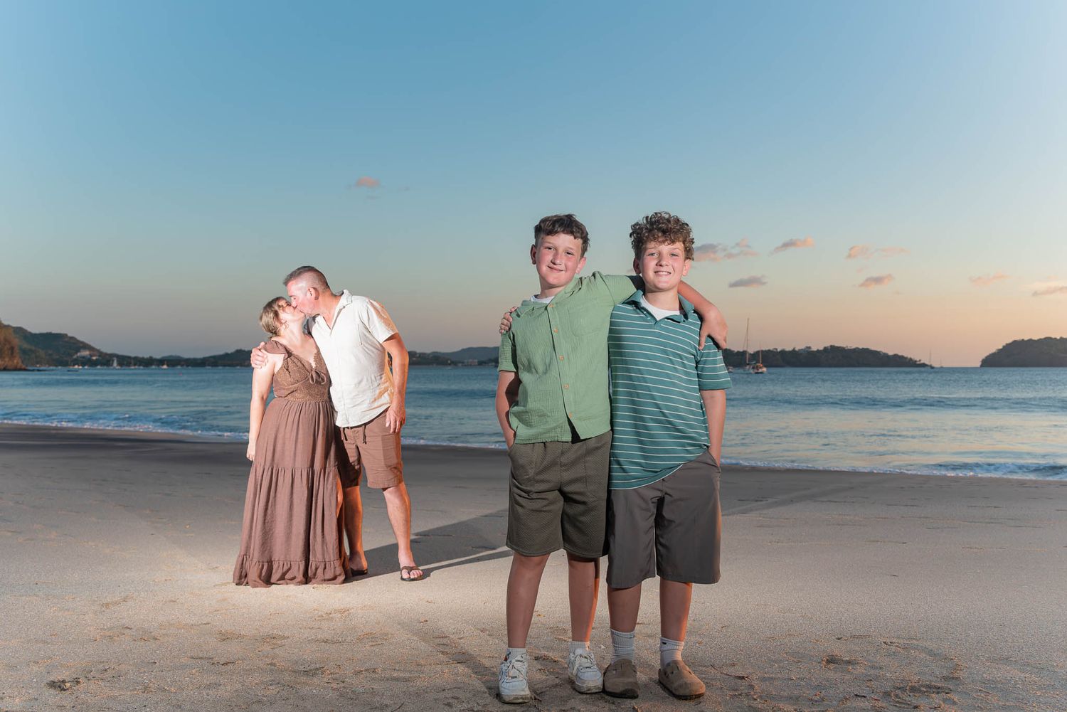 Family beach portrait at sunset with couple embracing and two boys smiling on sandy shore.