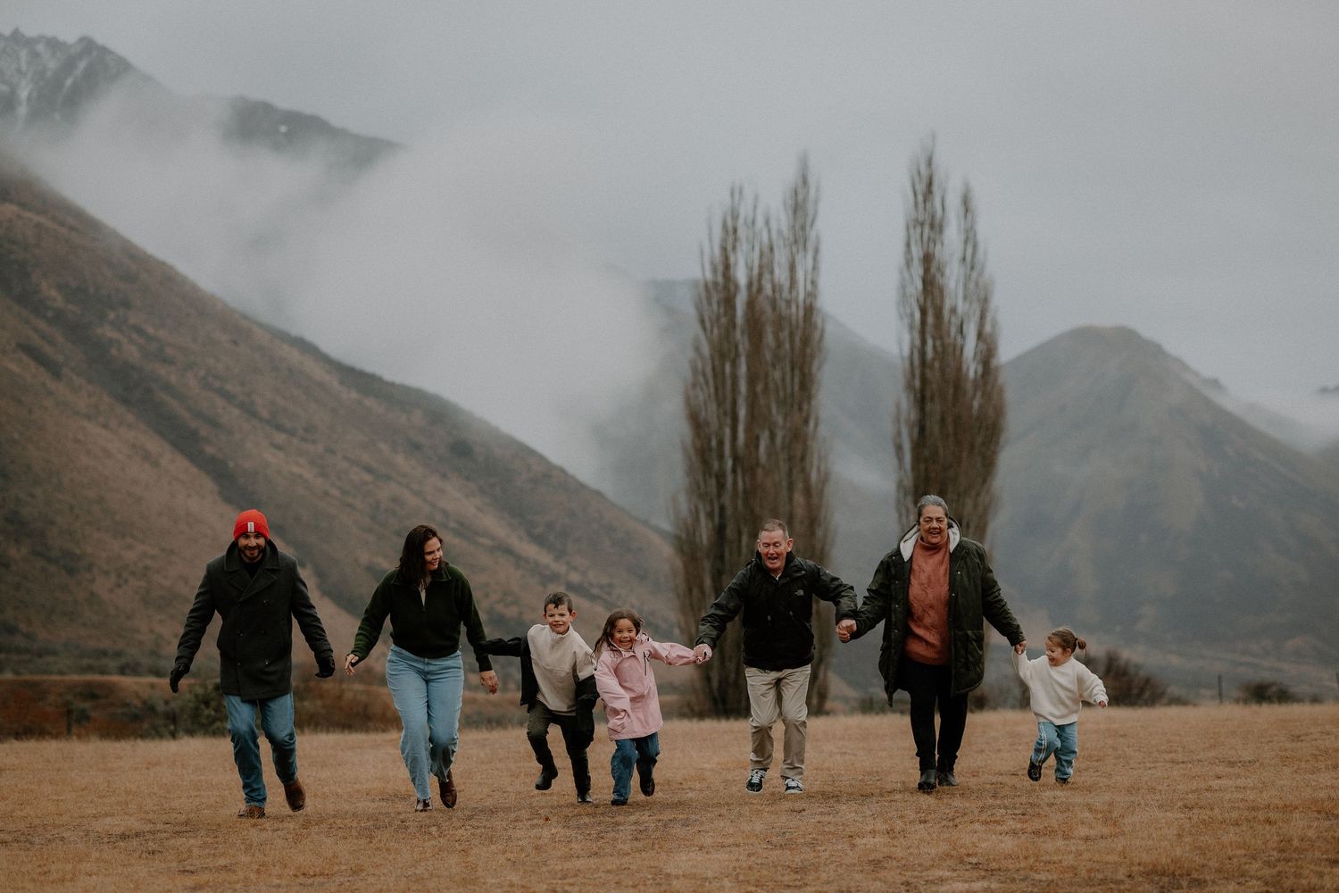 Family of seven holding hands running through misty mountain landscape on overcast day.