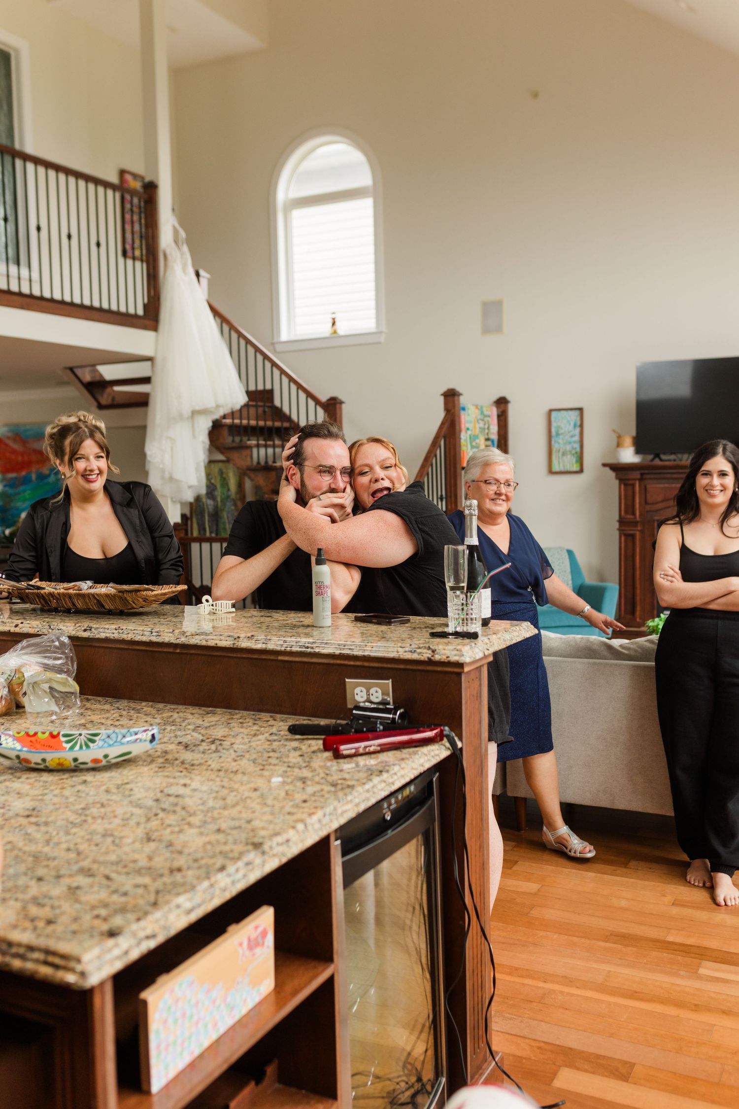 Group of people gathered in a home kitchen, smiling and hugging near a granite counter with a staircase in the background.