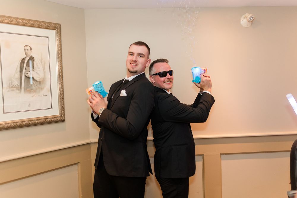 Two men in black suits pose back-to-back holding blue drinks at a formal event.