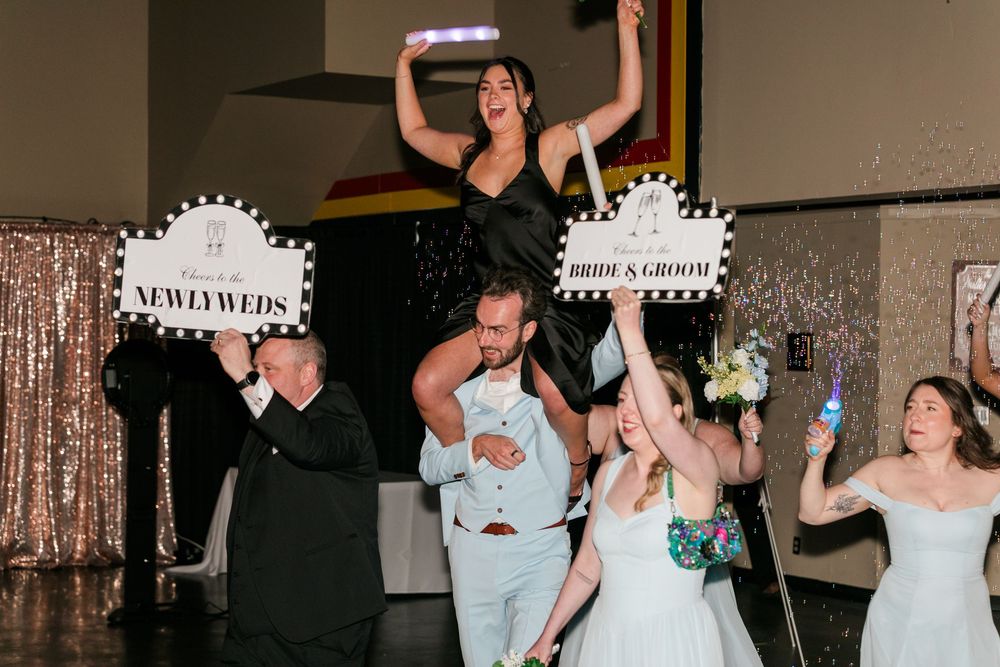 Newlyweds celebrate at wedding reception holding bride and groom signs with guests cheering and confetti flying.
