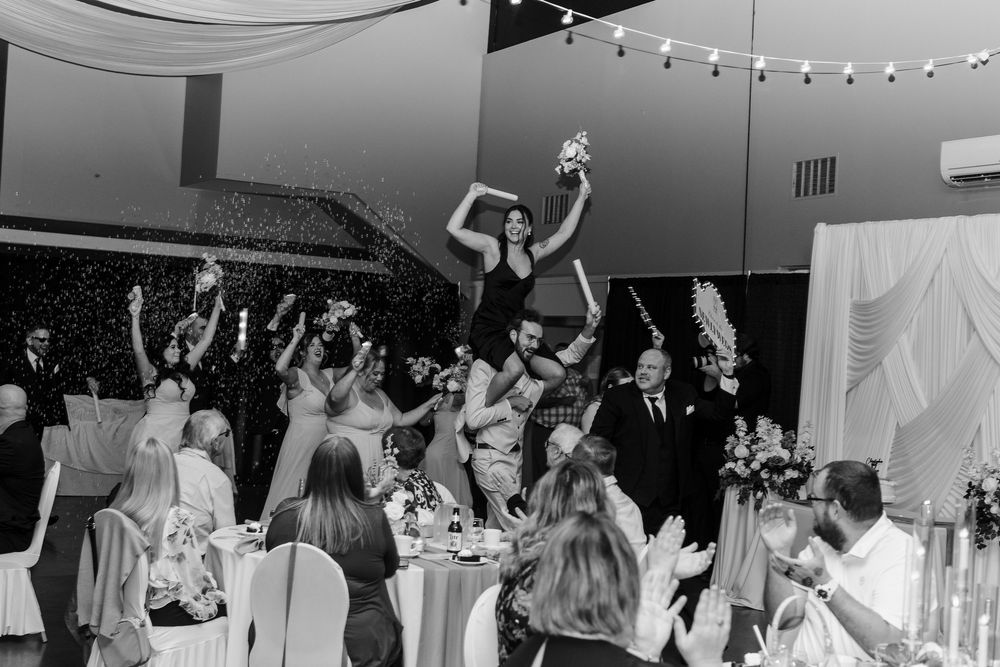 Black and white wedding reception photo of bride being lifted by guests while holding bouquet amid celebration.