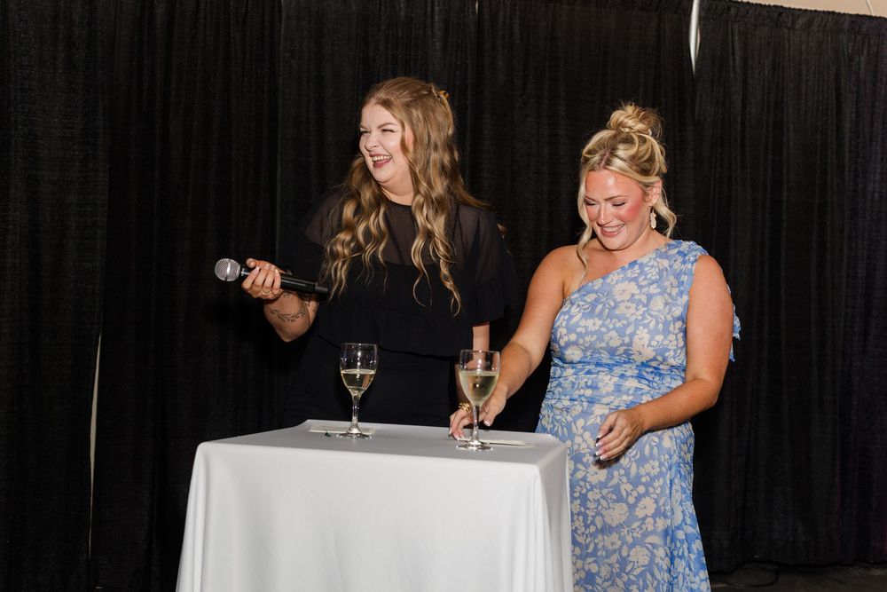 Two women laughing at a white table with champagne glasses, one holding a microphone at an elegant event.