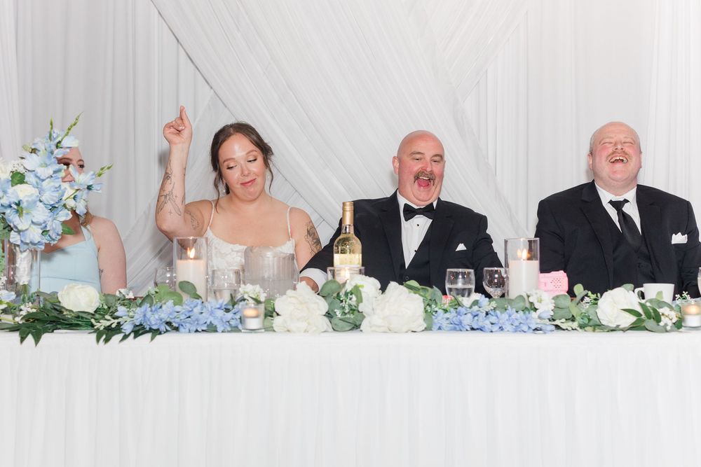 Bride and groom laugh with guest at elegant wedding reception head table adorned with blue and white floral arrangements.