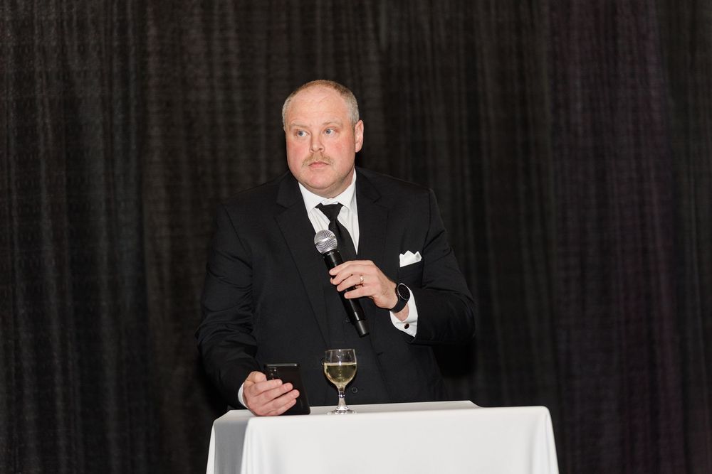 Man in black suit speaking at podium with microphone and white wine glass at formal event.