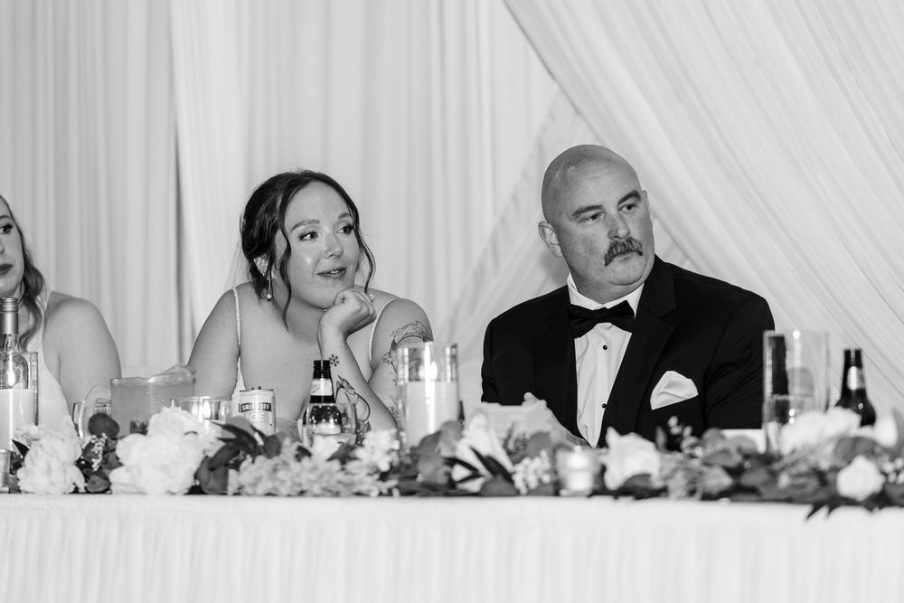Black and white photo of a bride and groom sitting at the head table during wedding reception speeches.